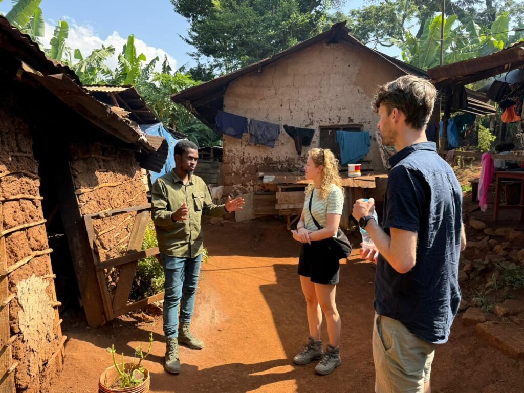 Three people stand in a sunny rural courtyard, talking near clay huts and hanging laundry between trees in a village setting.