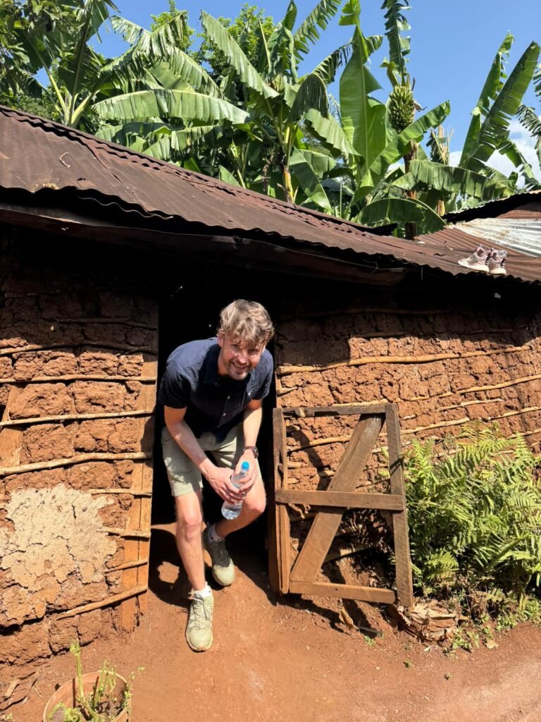 Man stepping out of a rustic mud‑brick hut, holding a water bottle, with banana plants and a blue sky behind.