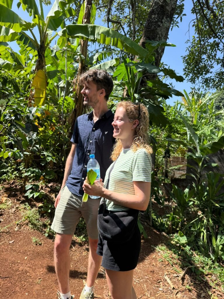 Two adults in a tropical garden, smiling at the camera among large banana plants to the left and right.