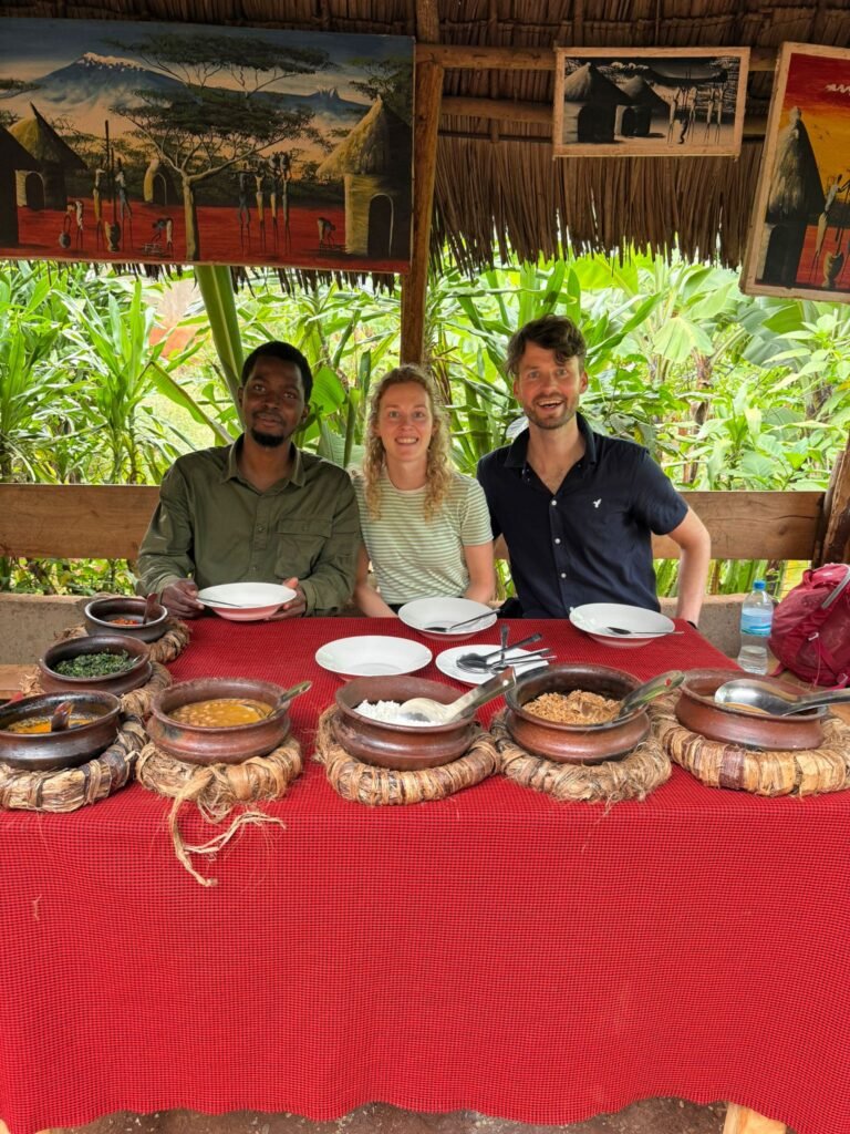 Three people sit side by side at a red tablecloth-covered table with clay bowls of food in front of them, smiling.