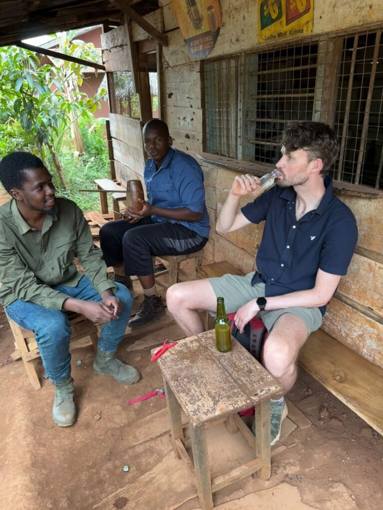 Three men sit on wooden benches outside a weathered building, sharing drinks.