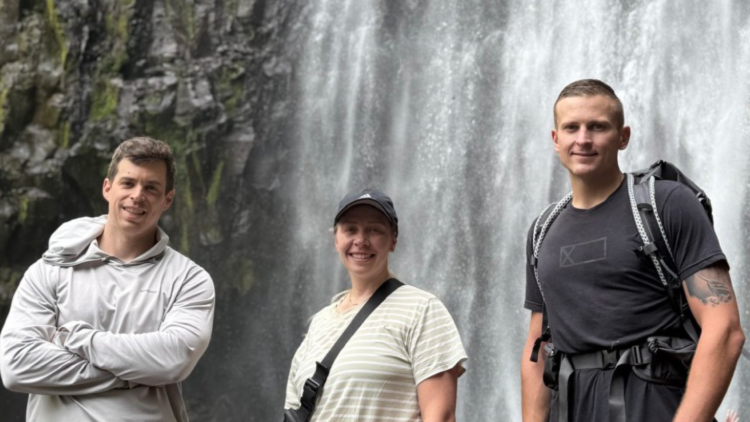 website | Chamecha Mana Culture Tourism Three hikers posing in front of a tall waterfall; a man in a gray hoodie, a woman in a striped shirt, and a man with a backpack smiling at the camera's inspiration behind them.